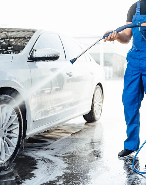 Professional washer in blue uniform washing luxury car with water gun on an open air car wash. Close up photo.