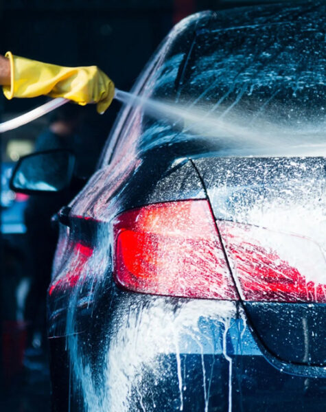 Professional washer in blue uniform washing luxury car with water gun on an open air car wash. Close up photo.